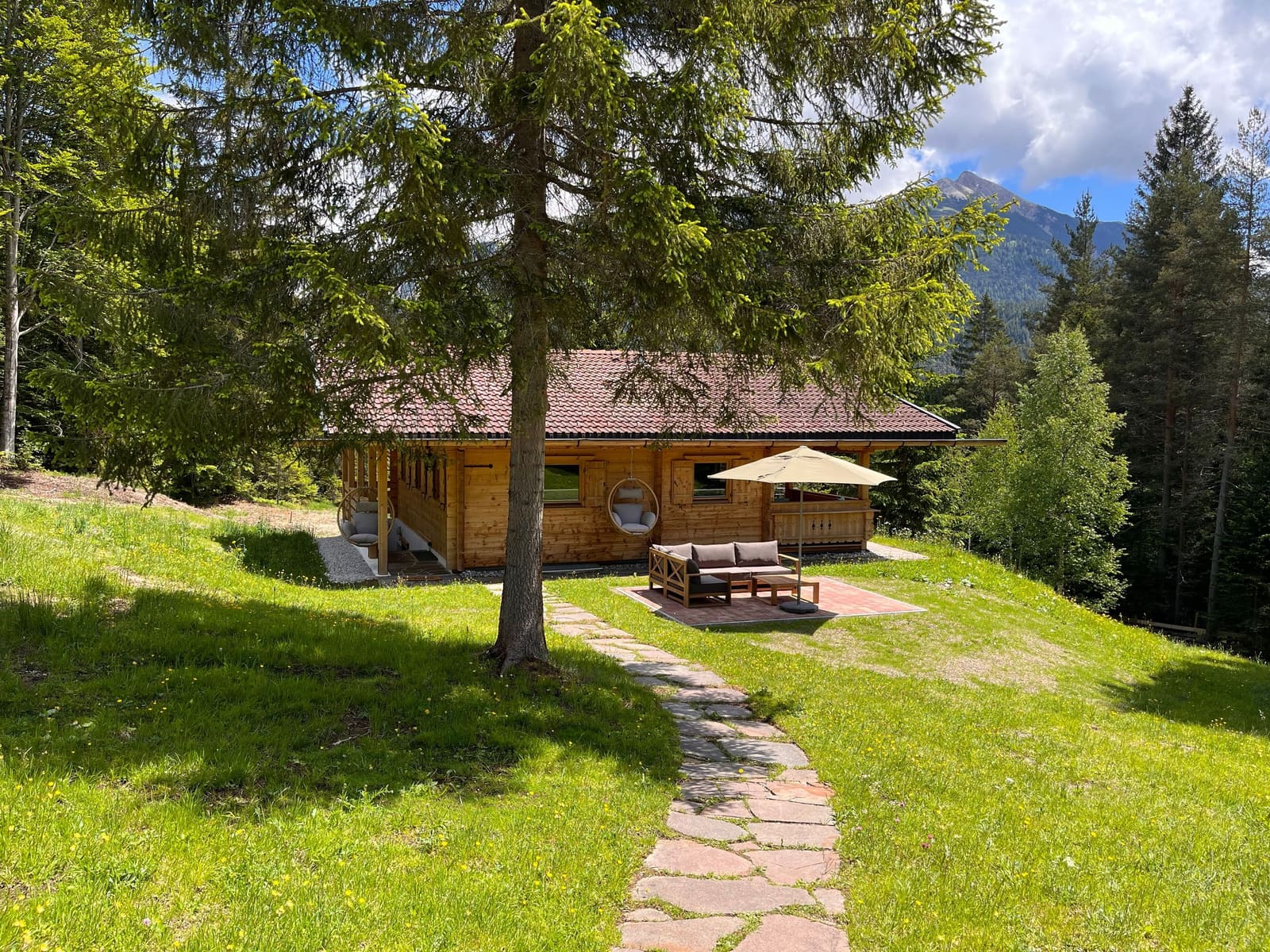 Das Chalet auf seiner Waldlichtung mit Steinweg, Hängesesseln und Bergpanorama im Sommer